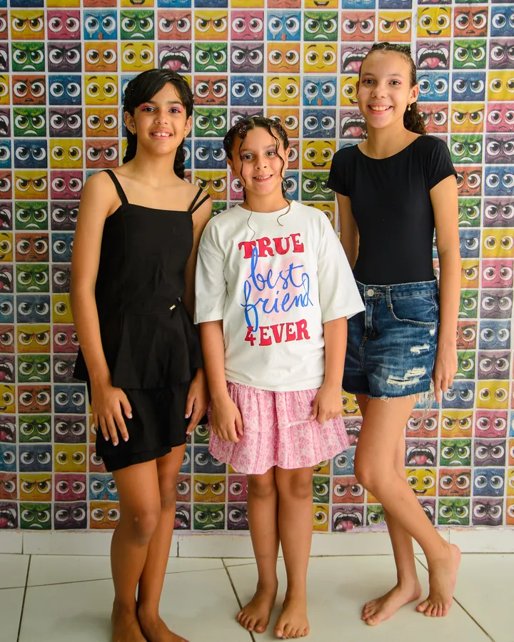 Três estudantes do Ensino Fundamental sorrindo juntas em frente a um painel colorido